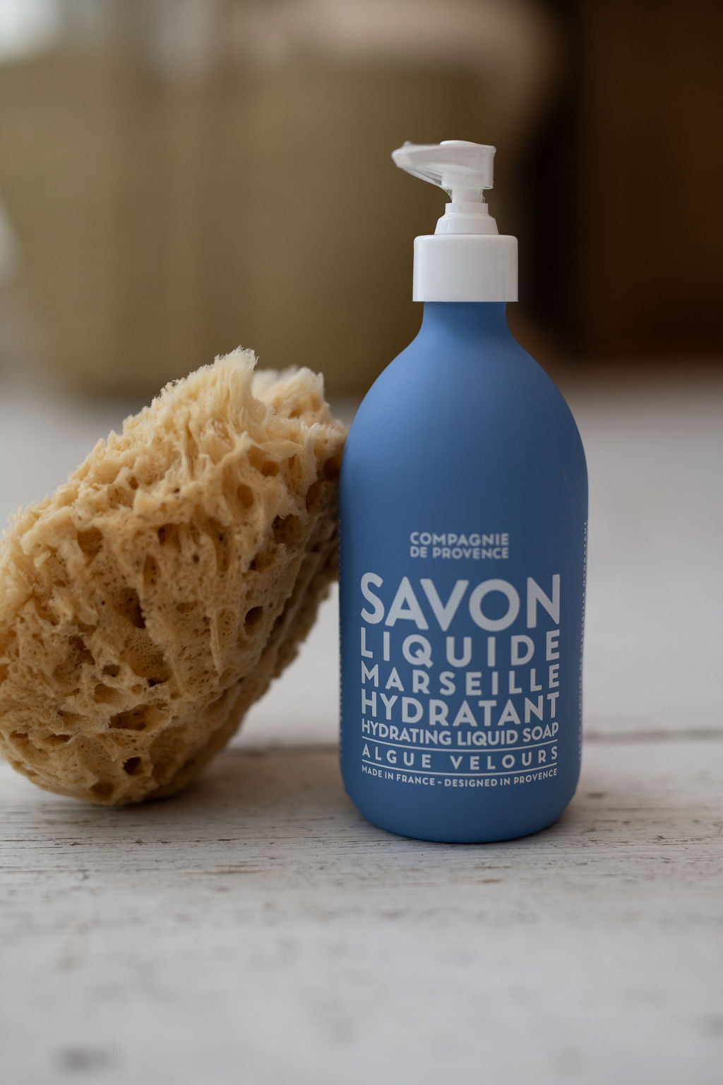 Blue bottle of Marseille liquid soap with a pump and a sponge on a wooden surface.