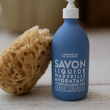 Blue bottle of Marseille liquid soap with a pump and a sponge on a wooden surface.